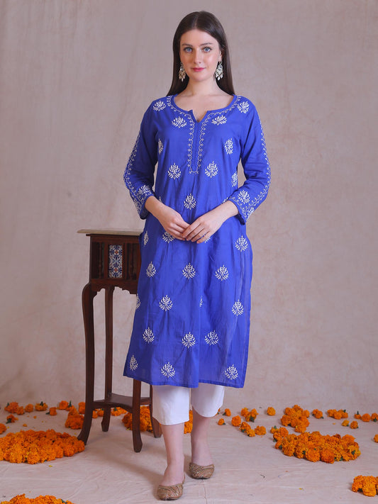Woman wearing a blue embroidered kurta with white pants standing in front of a beige wall with orange flowers on the floor.
