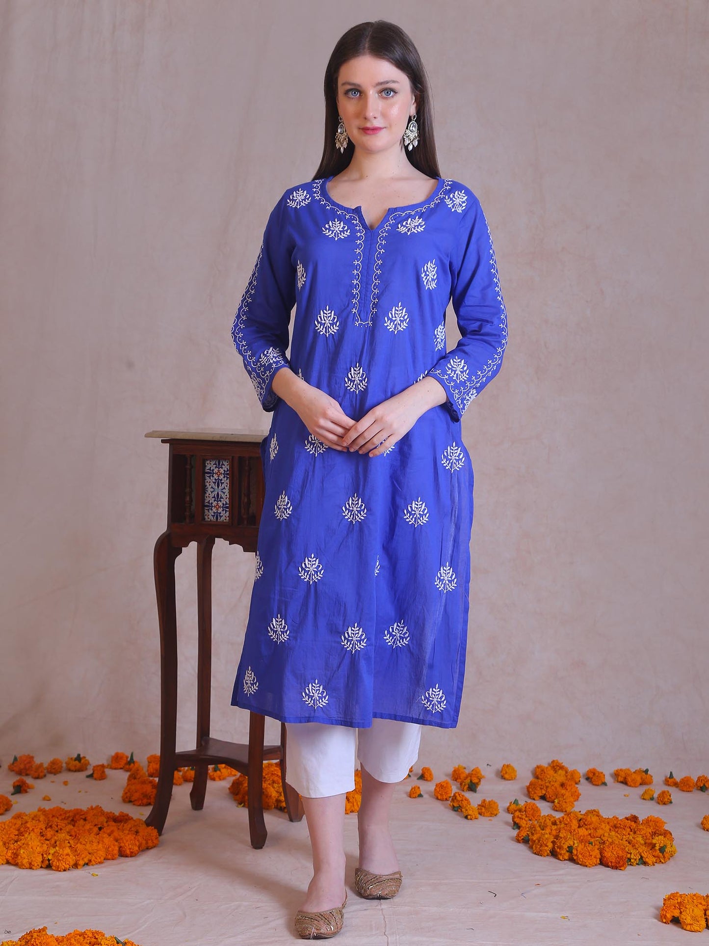 Woman wearing a blue embroidered kurta with white pants standing in front of a beige wall with orange flowers on the floor.