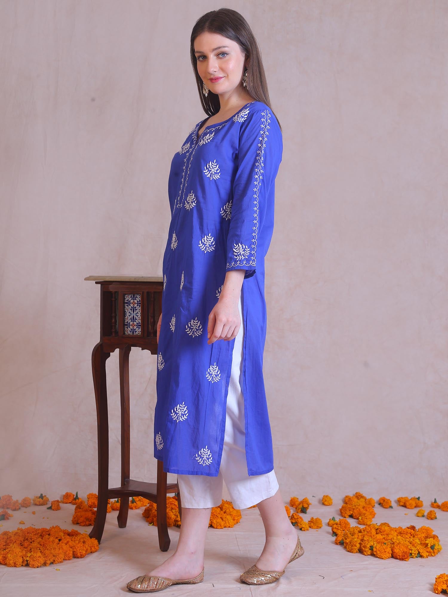 Woman wearing a blue embroidered kurta with white pants standing against a beige background with decorative elements.
